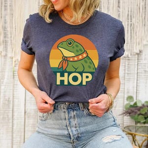 Puede incluir: Camiseta azul marino con una rana de dibujos animados con una bufanda roja y un collar con estrellas, sobre un diseño retro de puesta de sol. La palabra "HOP" está impresa en letras amarillas en negrita debajo de la rana.