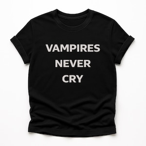 Puede incluir: Camiseta negra con el texto blanco "VAMPIRES NEVER CRY" en una fuente en negrita sin serifa. La camiseta tiene mangas cortas con puños enrollados. La tela parece ser una suave mezcla de algodón.