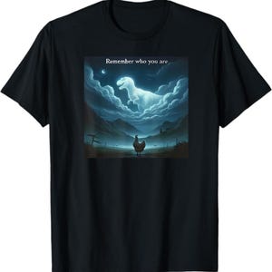 Puede incluir: Camiseta negra con un gráfico surrealista. El gráfico representa un pollo mirando hacia un T-Rex brillante en las nubes, con el texto "Remember who you are". El diseño está ambientado en un paisaje montañoso.