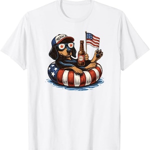 Könnte beinhalten: Weißes T-Shirt mit einem Cartoon-Dackel, der eine Sonnenbrille und eine Mütze mit der Aufschrift "AMERICA" trägt. Der Hund liegt in einem rot-weiß-blauen Schwimmring und hält eine Bierflasche und eine kleine amerikanische Flagge. Patriotisches Design.