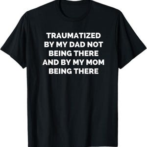 Puede incluir: Camiseta negra con texto blanco: "TRAUMATIZADO PORQUE MI PADRE NO ESTABA Y PORQUE MI MADRE SÍ ESTABA". La camiseta tiene cuello redondo y mangas cortas. El texto está centrado en la parte delantera.