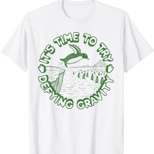 Könnte beinhalten: Weißes T-Shirt mit einer grünen Grafik eines Pinguins, der von einer Klippe springt, mit dem Text "IT'S TIME TO TRY DEFYING GRAVITY" in einem kreisförmigen Design. Das Design beinhaltet weitere Pinguine und eine Bergkette.