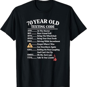 Puede incluir: Camiseta negra con texto blanco "70 YEAR OLD TEXTING CODE" y una lista de acrónimos humorísticos. Incluye un pequeño gráfico de una persona cayendo y una llama.