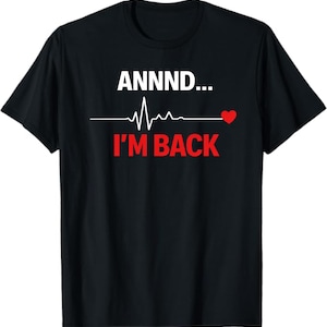 Op de afbeelding: Zwart t-shirt met de tekst "ANNNND... I'M BACK" in wit en rood. Een witte hartslaglijn grafiek staat tussen de tekst, met een rood hart aan het einde. Het t-shirt heeft een ronde hals.