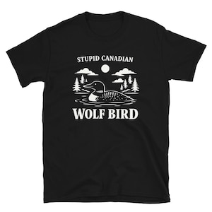 Pode incluir: Camiseta preta com um gráfico branco de um mergulhão na água, rodeado por árvores, nuvens e uma lua. O texto acima diz "STUPID CANADIAN" e abaixo "WOLF BIRD".