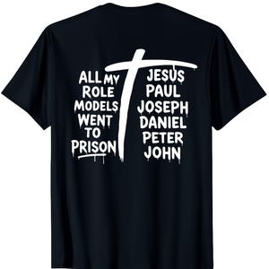 May include: Black t-shirt with a white cross and text. The text on the left reads "ALL MY ROLE MODELS WENT TO PRISON". The text on the right reads "JESUS PAUL JOSEPH DANIEL PETER JOHN".