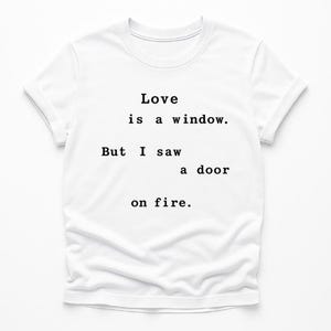 Op de afbeelding: Wit t-shirt met de zwarte tekst "Love is a window. But I saw a door on fire." gecentreerd op de voorkant. Het shirt heeft korte mouwen en een ronde hals.