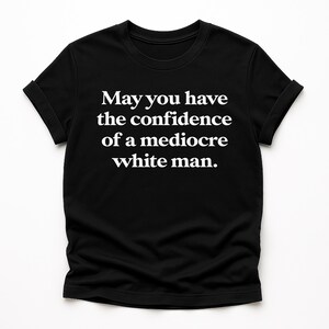 Op de afbeelding: Zwart T-shirt met de witte tekst: "Moge je het zelfvertrouwen hebben van een middelmatige blanke man." Het shirt heeft een ronde hals en korte mouwen met opgerolde manchetten. Het shirt wordt op een witte achtergrond gepresenteerd.