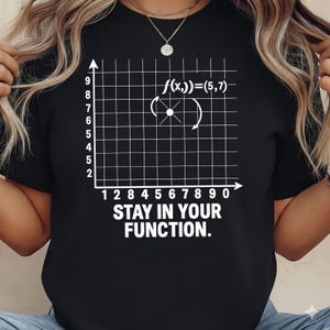May include: Black t-shirt featuring a white graph with the equation f(x,))=(5,7) and the text "STAY IN YOUR FUNCTION." The shirt has a crew neck and short sleeves.