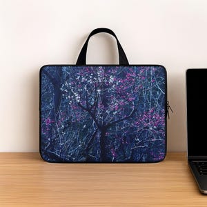 May include: A rectangular laptop sleeve featuring a dark, moody forest scene with bare branches and clusters of white and magenta blossoms. The sleeve has a black top handle, a black zipper closure, and a slim, structured profile for device storage.