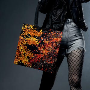 Könnte beinhalten: Schwarze Tragetasche mit einem lebendigen Herbstlaub-Design in Orange und Gelb auf dunklem Hintergrund. Die Tasche hat einen schwarzen Riemen. Die Tasche wird von einer Person getragen, die eine schwarze Jacke und Jeansshorts trägt.