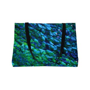 May include: A tote bag featuring a vibrant, abstract design of overlapping blue and green leaves. The bag has black straps and a white background. The overall design is reminiscent of a lush, natural landscape.