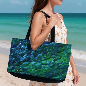May include: A large tote bag with a black base and straps. The bag features a vibrant, abstract pattern of blue and green leaf-like shapes. The bag is being carried on a beach with turquoise water in the background.