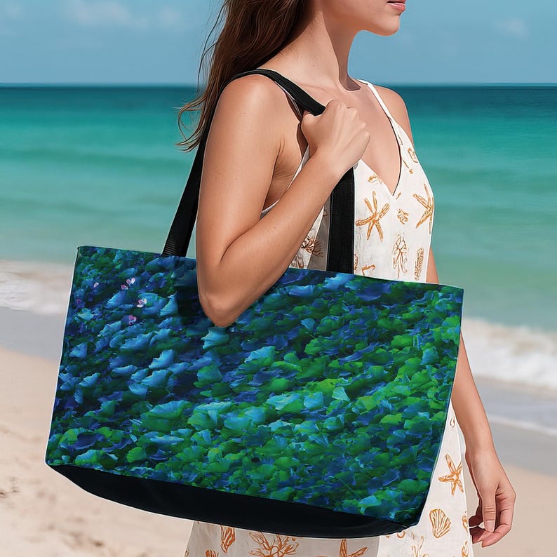 May include: A large tote bag with a black base and straps. The bag features a vibrant, abstract pattern of blue and green leaf-like shapes. The bag is being carried on a beach with turquoise water in the background.