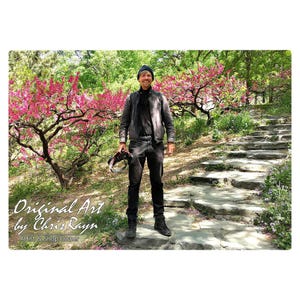 May include: A person stands on stone steps in a park, holding a camera. The person wears a black jacket, pants, and a beanie. Pink flowering trees and green foliage frame the scene. Text reads "Original Art by Chris Rayn, Artist & Shop Owner."