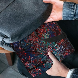 Puede incluir: Una funda negra para port&aacute;til con un dise&ntilde;o colorido de ramas de &aacute;rboles se coloca en una mochila gris. La funda presenta un asa negra y un patr&oacute;n vibrante de hojas rojas sobre un fondo azul oscuro y verde.