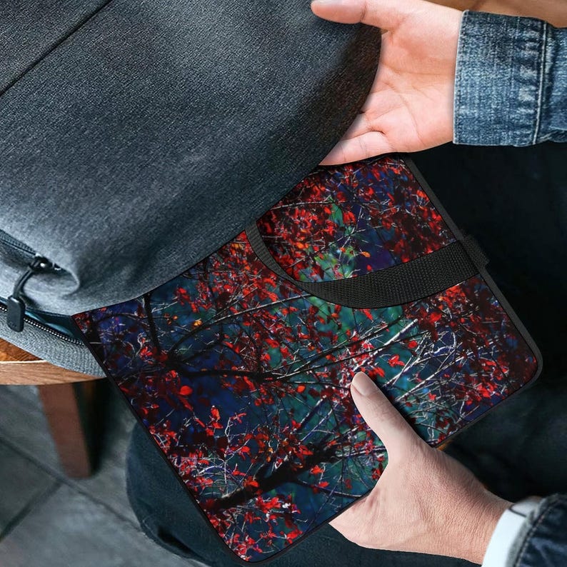 Puede incluir: Una funda negra para port&aacute;til con un dise&ntilde;o colorido de ramas de &aacute;rboles se coloca en una mochila gris. La funda presenta un asa negra y un patr&oacute;n vibrante de hojas rojas sobre un fondo azul oscuro y verde.