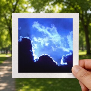 May include: A square photographic print featuring a dramatic sky with deep blue tones and bright white clouds. The lower portion of the image shows dark, silhouetted cloud formations with light glowing along the edges against a vibrant blue sky.
