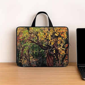 May include: A rectangular laptop sleeve featuring a photographic print of a dense forest scene during autumn. The design displays a large tree trunk with dark bark surrounded by foliage in vibrant shades of yellow, orange, and green leaves.