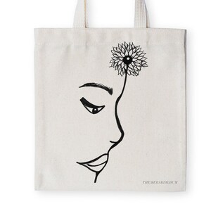 May include: A white canvas tote bag with a black line drawing of a woman's face with a flower on her head. The text "THEMERAKIALBUM" is printed on the bottom of the bag.