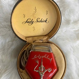 May include: Vintage Lady Schick electric razor in a round, black case. The red razor has a silver design and the words "Lady Schick" in silver script. The case interior is tan with the words "Lady Schick" in black script.