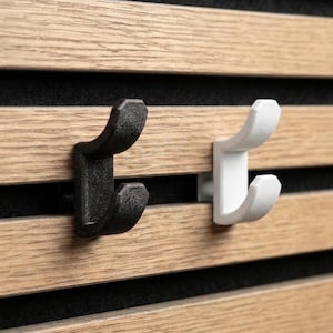 Könnte beinhalten: Zwei Wandhaken, einer schwarz und einer weiß, an einer Holzlattenwand befestigt. Die Haken haben ein gebogenes Design, das zum Aufhängen von Gegenständen geeignet ist. Die Holzlatten sind hellbraun mit schwarzen Zwischenräumen.