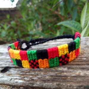 May include: Handmade woven bracelet featuring a vibrant pattern of red, yellow, green, and black squares. The bracelet is made of colorful threads and has a black cord closure. The design is reminiscent of a flag.