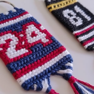 May include: Two handmade keyrings with woven designs. One is blue, red, and white with the number "24" in white. The other is black, white, yellow, and red with the number "81" in white. Both have tassels.