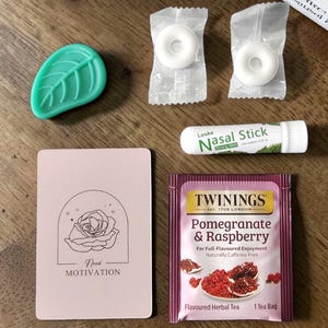 May include: An assortment of items on a wooden surface. Includes a green leaf-shaped object, two wrapped mints, a nasal stick, a pink card with a rose illustration and the words "Need Motivation", and a Twinings Pomegranate & Raspberry tea bag.