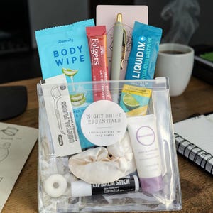 May include: A clear plastic pouch labeled Night Shift Essentials containing a body wipe, a Folgers coffee stick, a pen, a Liquid I.V. packet, dental floss, a white scrunchie, a tube of hand lotion, and a green tea lip balm stick.