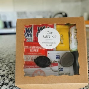 May include: A brown cardboard box with a scalloped window containing a car care kit. The kit includes stain removal wipes, a yellow towel packet, Zeiss glasses wipes, an emergency medical card, and a black key fob with a metal ring.
