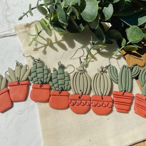 May include: A set of eight handmade polymer clay earrings featuring different succulent and cactus designs. The earrings are painted in shades of green and orange and have silver hooks.