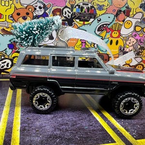 May include: A gray and black Jeep toy car with a small green Christmas tree on top. The toy car has black wheels and red accents. The background is a colorful collage of stickers.