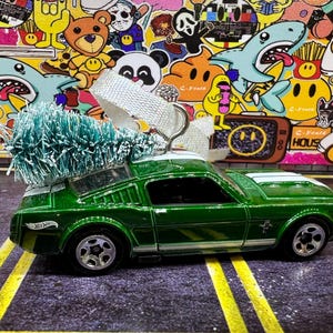 May include: A green toy car with a white stripe on the hood and a small Christmas tree on top. The car has black windows and silver wheels. The background is a colorful collage of stickers.