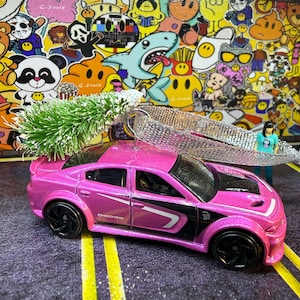 May include: A pink Dodge Charger toy car with black accents and wheels. The car has a white stripe design and the word "Dodge" is visible. A small green tree and silver ribbon are attached to the car.