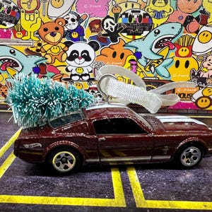 May include: A burgundy die-cast model car with white racing stripes and a small green Christmas tree on top. The car has a white ribbon for hanging. The background is a colorful collage of cartoon characters.