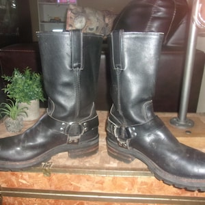 May include: A pair of black leather motorcycle boots with a square toe and a harness strap. The boots have a mid-calf height and a rugged, worn appearance. The soles are thick and the heels are low.