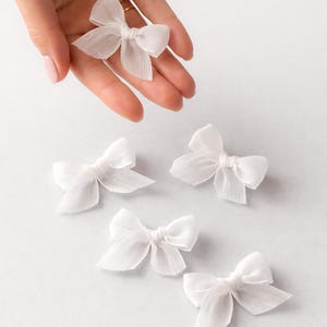 Könnte beinhalten: Mehrere kleine, weiße Stoffschleifen mit durchscheinender Textur werden vor einem hellen Hintergrund gezeigt. Eine Schleife wird von einer Hand gehalten, andere sind darunter verstreut. Diese zarten Schleifen eignen sich zum Basteln oder Dekorieren.