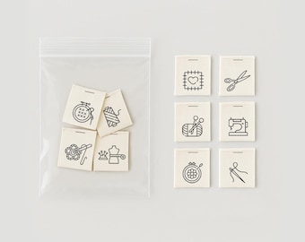 May include: A set of eight white fabric labels with black line drawings of sewing and crafting tools. The labels include a sewing machine, scissors, a needle and thread, a spool of thread, a button, a thimble, a heart, and a hoop with cross-stitches.