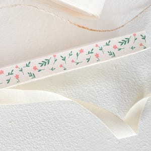 May include: Close-up of a cream-colored ribbon with a floral pattern. The ribbon features small pink flowers and green leaves. A roll of ribbon and a loose ribbon are also visible.