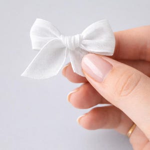 Può includere: Un piccolo fiocco di tessuto bianco con un centro annodato e code fluenti. Il fiocco è tenuto in mano, mostrando la sua delicata consistenza e il suo design semplice. Perfetto per progetti di artigianato o abbellimento.