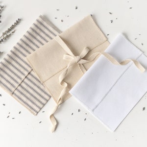 Puede incluir: Tres sobres de tela en beige, blanco y con patrones de rayas. El sobre beige está atado con una cinta color crema. Ramas de lavanda secas y capullos sueltos están esparcidos alrededor de los sobres sobre una superficie blanca.