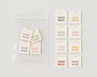 May include: A collection of off-white fabric tags with the words "HAND MADE" printed in various colors, including purple, green, and pink. Some tags are in a clear plastic bag, while others are arranged on a light gray background.