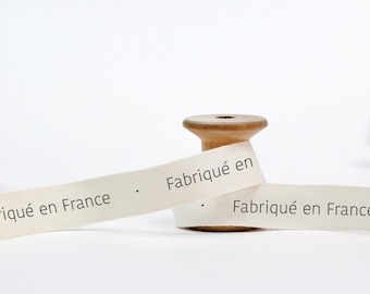May include: A spool of white cotton ribbon with the text "Fabriqué en France" printed on it. The ribbon is wrapped around a wooden spool.