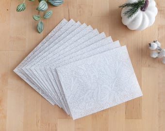May include: A stack of light gray envelopes with a white floral pattern. The envelopes are fanned out on a wooden surface, with decorative elements including a white pumpkin and sprigs of greenery.
