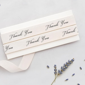 Puede incluir: Una pila de tarjetas blancas con cintas color crema. Las cintas llevan la inscripción "Thank You" en elegante escritura negra. Una ramita de lavanda y pétalos sueltos están esparcidos alrededor de las tarjetas sobre una superficie gris claro.