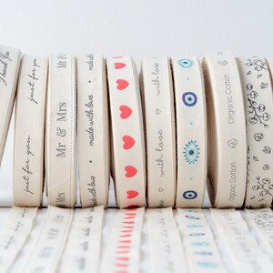 May include: A collection of beige ribbon rolls with various printed designs and text. The ribbons feature phrases like "Thank You," "just for you," and "Mr & Mrs," along with heart and eye patterns. The ribbons are made of organic cotton.