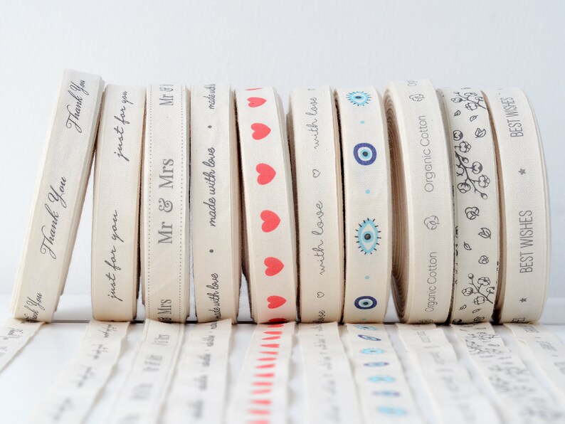 May include: A collection of beige ribbon rolls with various printed designs and text. The ribbons feature phrases like "Thank You," "just for you," and "Mr & Mrs," along with heart and eye patterns. The ribbons are made of organic cotton.