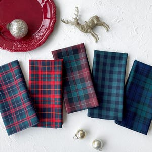 May include: Five folded fabric napkins featuring various plaid patterns in red, green, and blue colorways. A red plate, a silver reindeer ornament, and two small silver spherical ornaments are arranged around the napkins on a white surface.