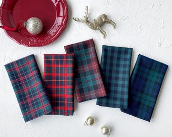 May include: Five folded fabric napkins featuring various plaid patterns in red, green, and blue colorways. A red plate, a silver reindeer ornament, and two small silver spherical ornaments are arranged around the napkins on a white surface.